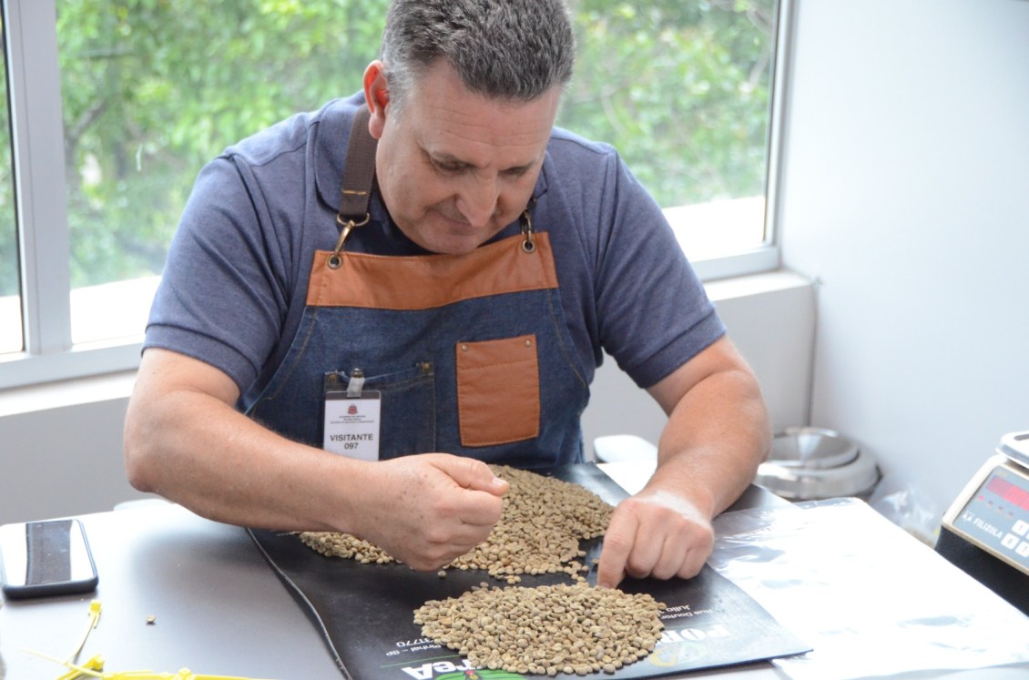 Homem examinando grãos de café em uma mesa, com amostras espalhadas à sua frente e um ambiente iluminado ao fundo.