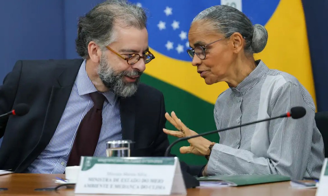 Dois ministros conversam em uma mesa, com a bandeira do Brasil ao fundo.
