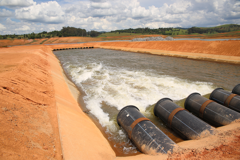 Vista de um canal de água com tubos de PVC preto na borda, cercado por terra vermelha e vegetação ao fundo sob um céu nublado.