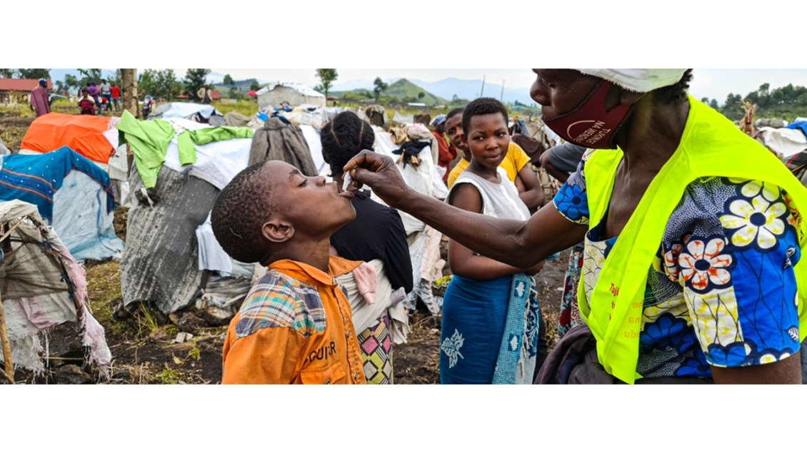Uma mulher administrando a vacina oral contra a cólera a uma criança em um campo de refugiados, com outras pessoas observando ao fundo.
