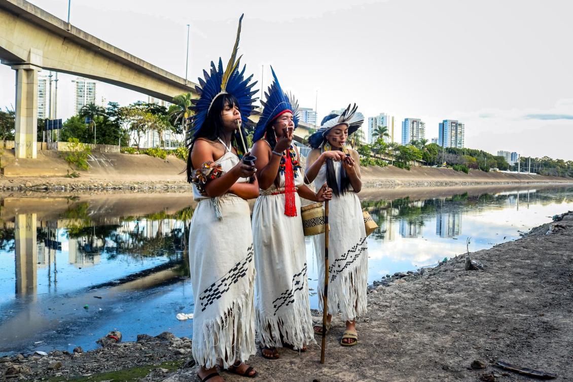 Três mulheres vestindo roupas tradicionais e adornos indígenas, em frente a um rio, com um cenário urbano ao fundo.