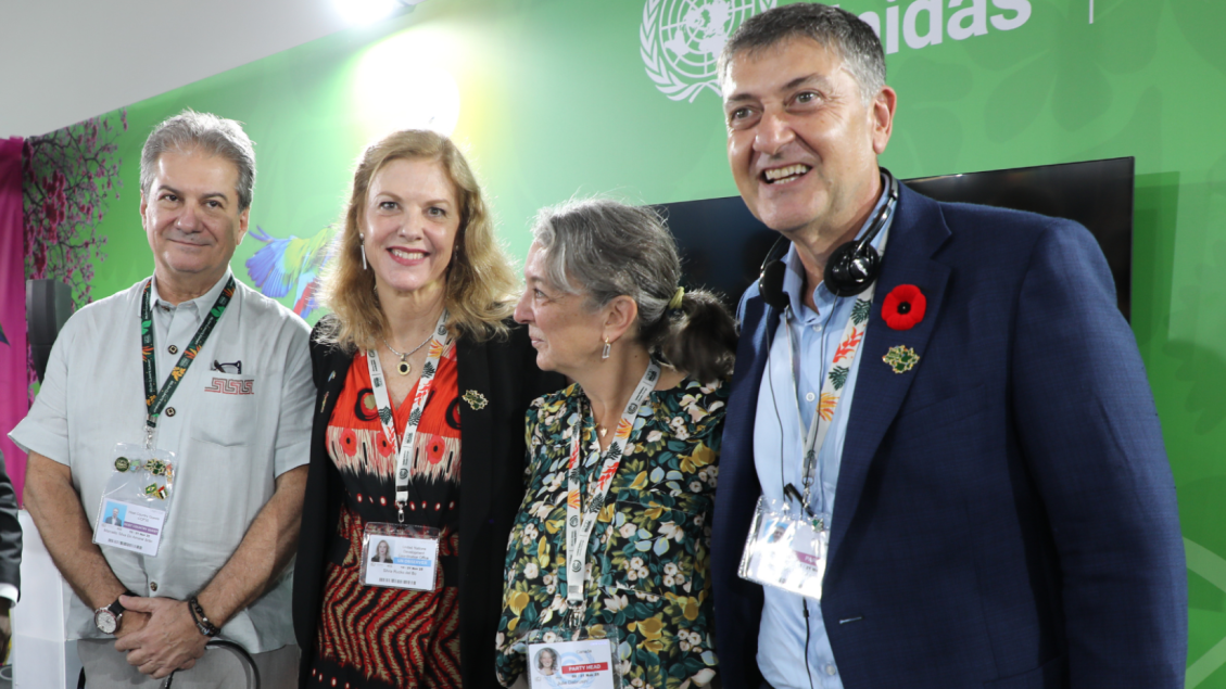Quatro profissionais sorrindo em um evento da ONU, com fundo verde e logotipo da ONU visível.