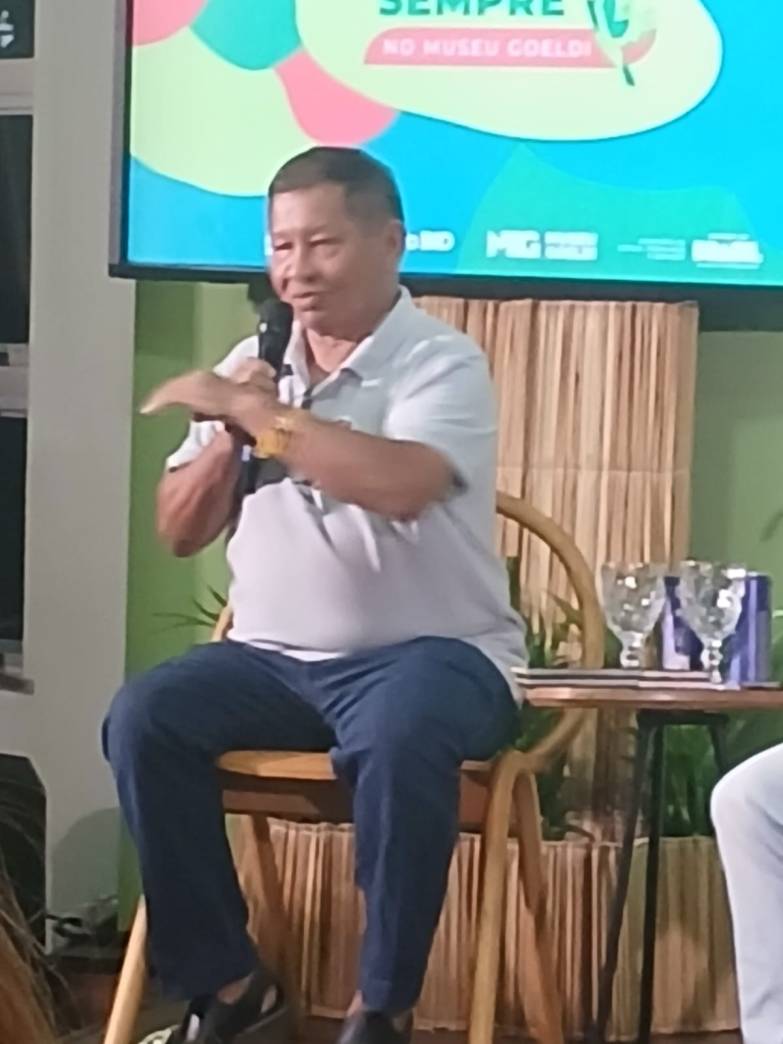 Homem em camisa clara falando ao microfone em um evento no Museu Goeldi, com um fundo colorido e decoração natural.