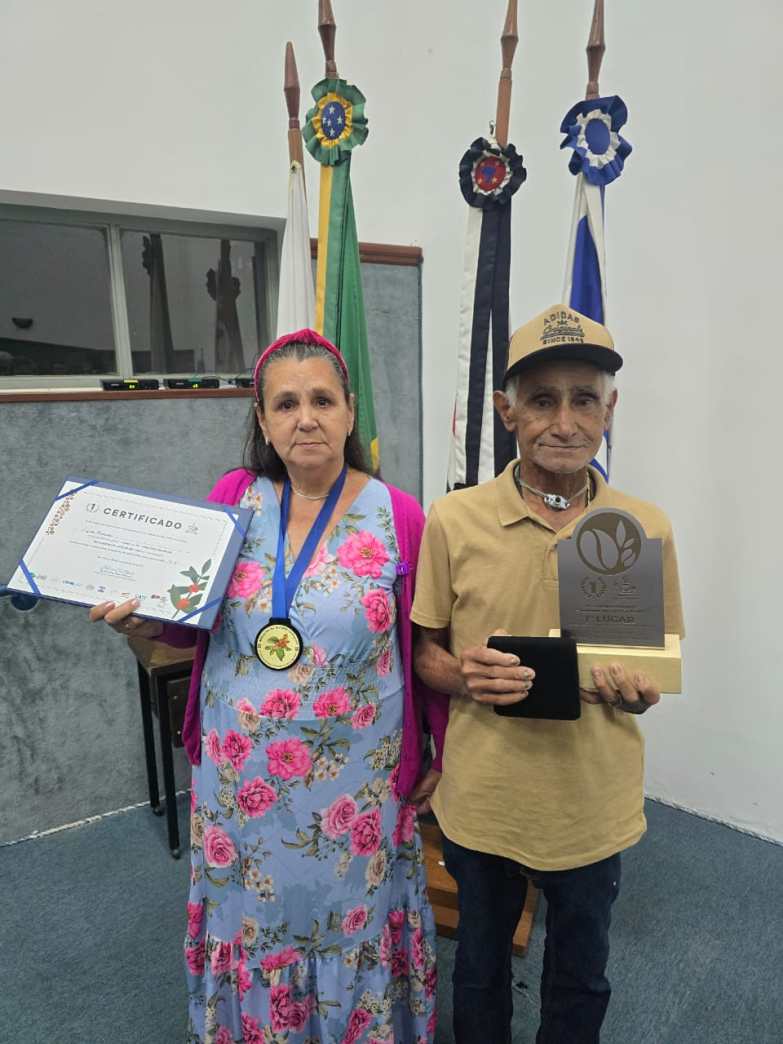 Casal posando em uma cerimônia de premiação, segurando um certificado e um troféu, com bandeiras ao fundo.