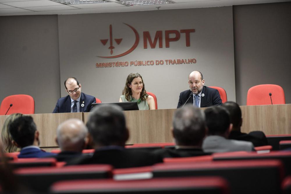 Reunião no Ministério Público do Trabalho (MPT) com três pessoas em destaque, um homem de óculos, uma mulher e um homem com terno, discutindo sobre segurança no trabalho.